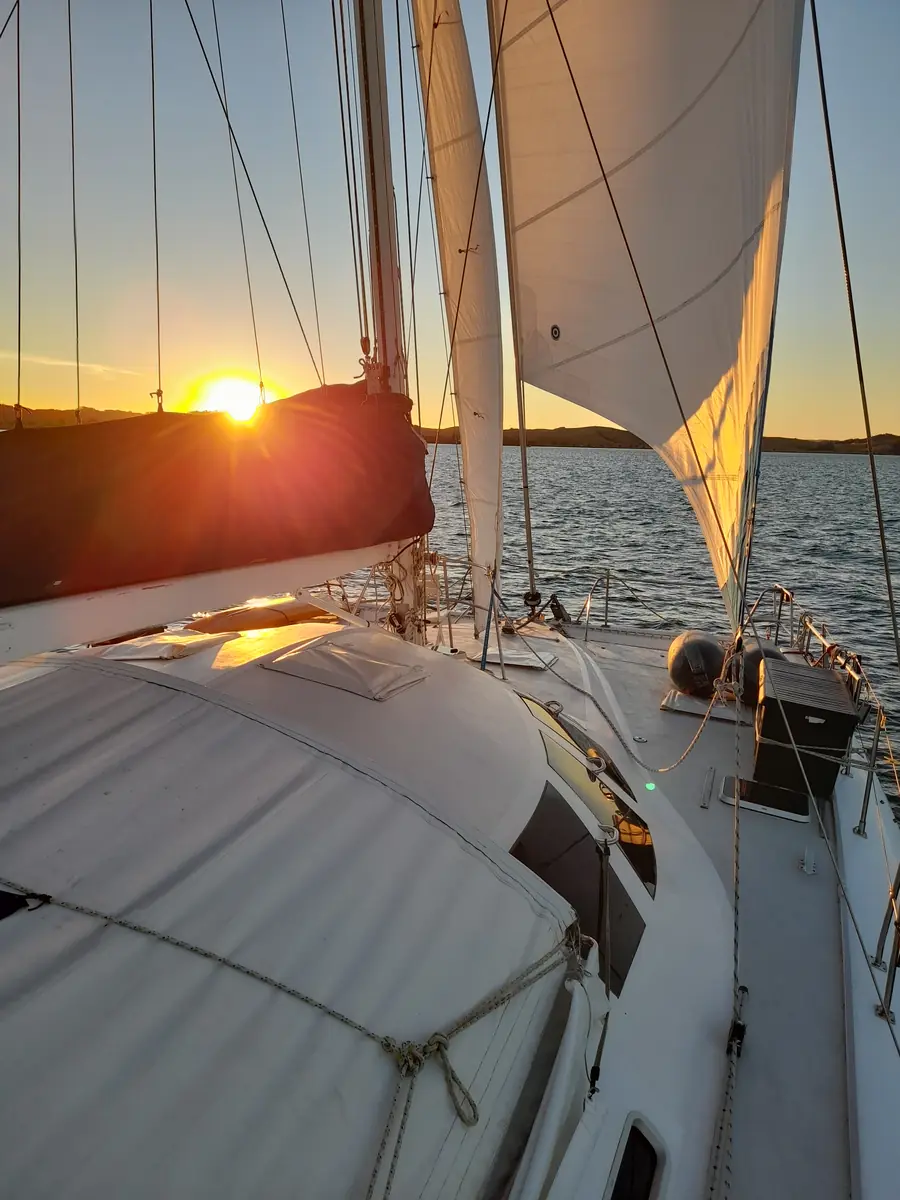 Looking along the deck of a sailing yacht towards the setting sun, sails above