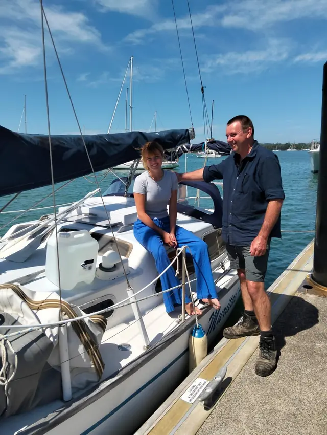 A young girl sitting in the cockpit of a small sailboat at a dock, with her dad standing beside it