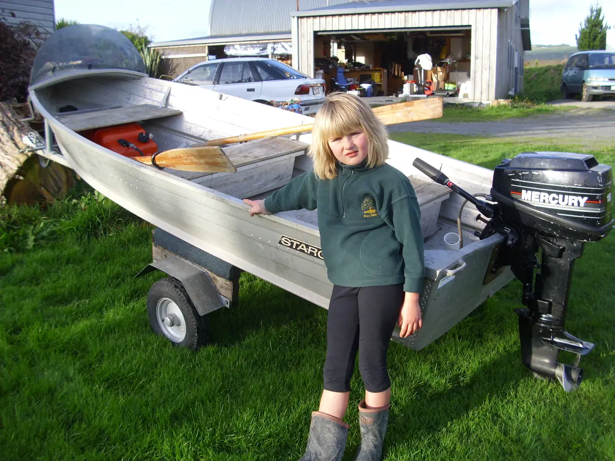 A young girl in gumboots standing proudly beside a small aluminium dinghy on a trailer, with an outboard motor